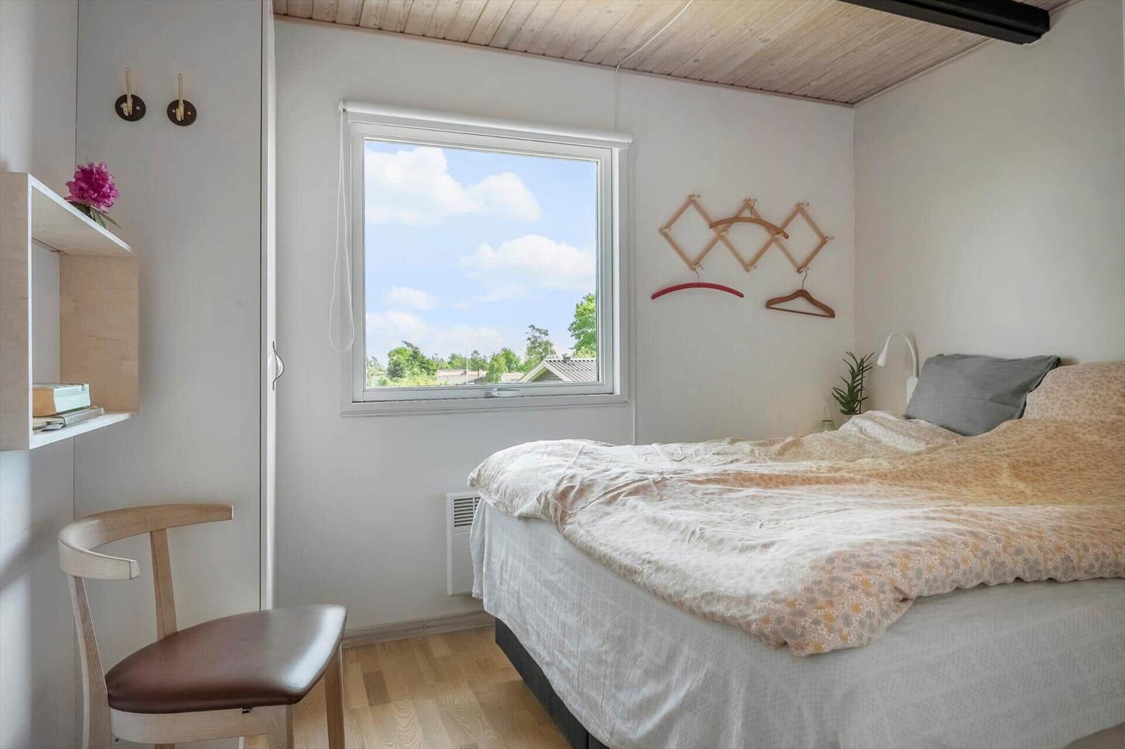 Schlafzimmer mit Bett, Fenster und Stuhl. Holzdecke und Wandregal.