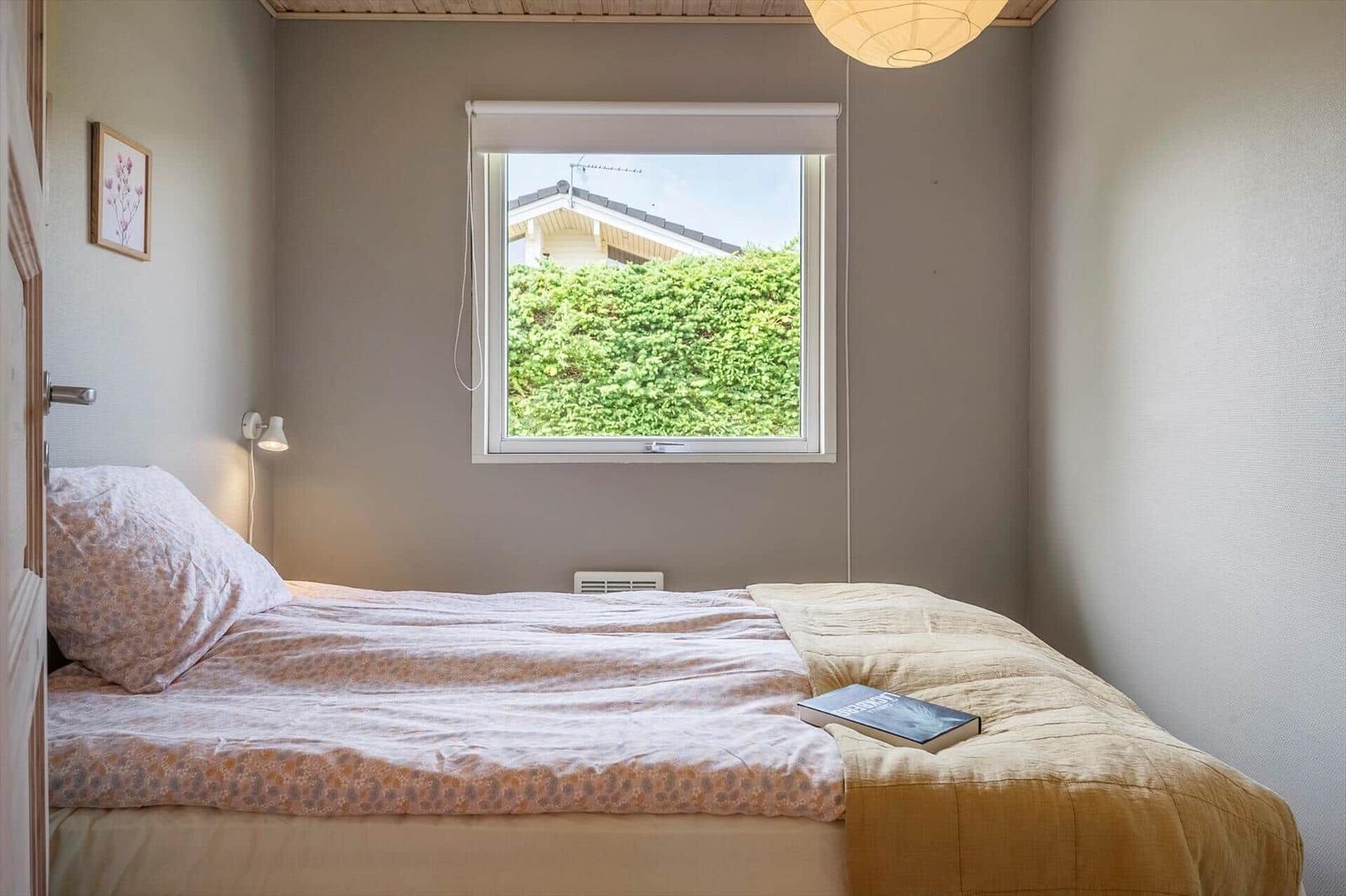 Schlafzimmer mit Bett, Fenster und Lampe. Draußen grüne Pflanzen und Hausdach.