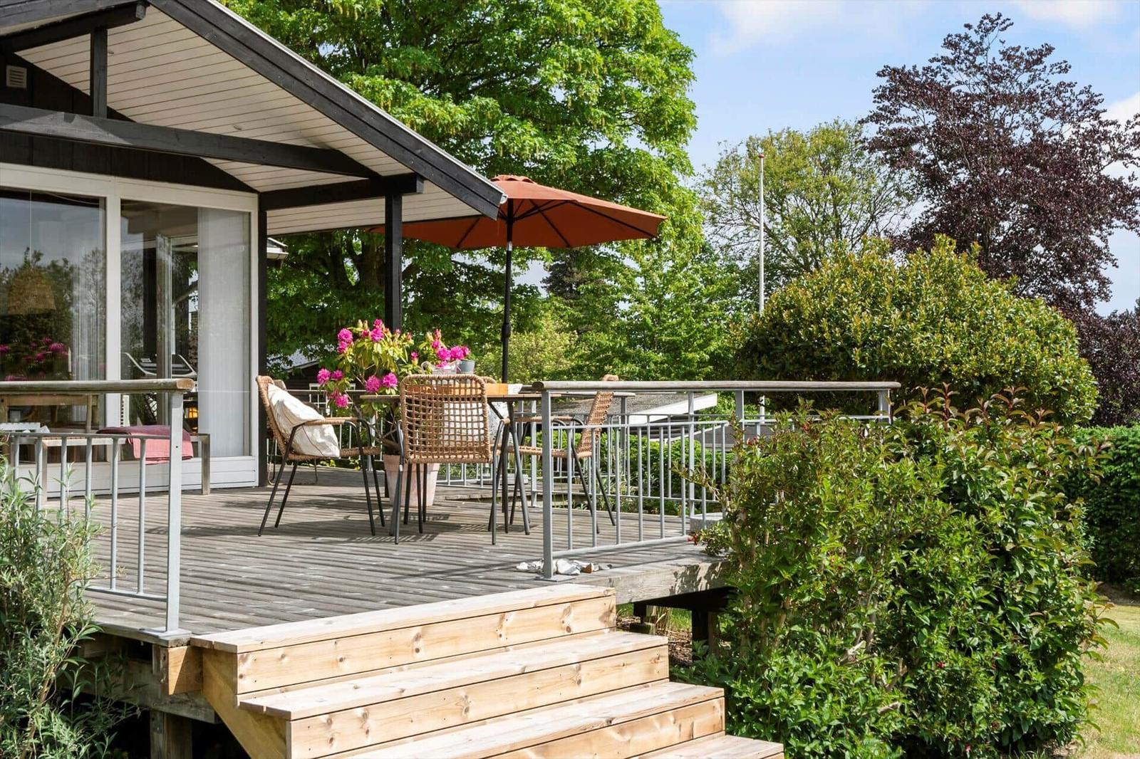 Terrasse mit Tisch, Stühlen und Sonnenschirm. Umgeben von Grün und Blumen.