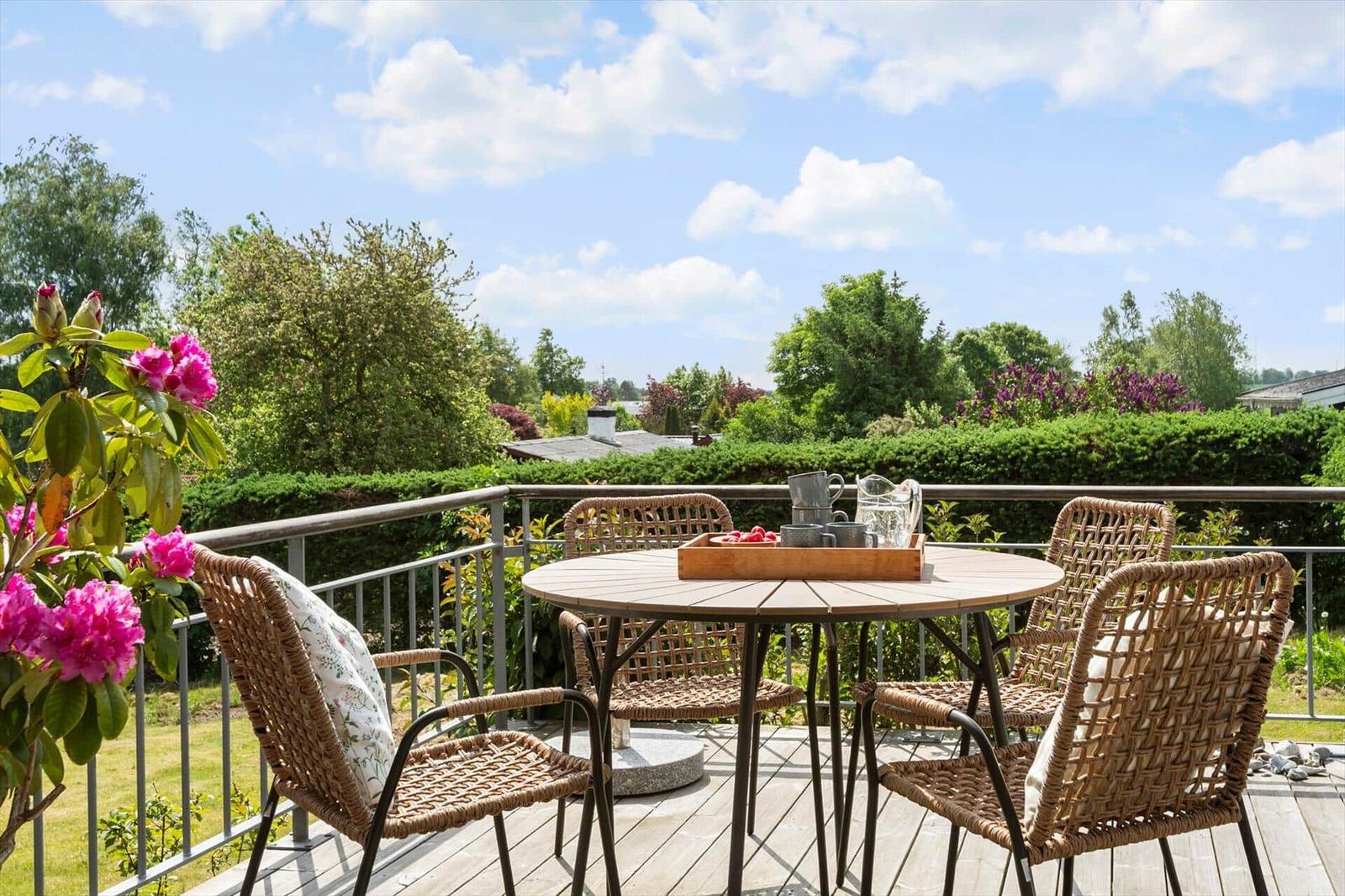 Terrasse mit Tisch und Stühlen, Blick auf grüne Bäume und Sträucher unter blauem Himmel.