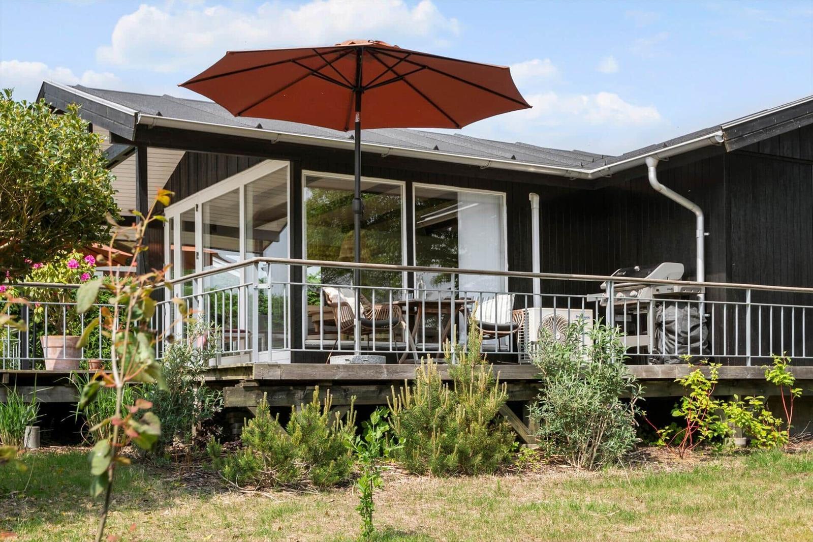 Terrasse mit Sonnenschirm, Grill und Sitzgelegenheiten. Dunkelholzhaus mit Glasfront.