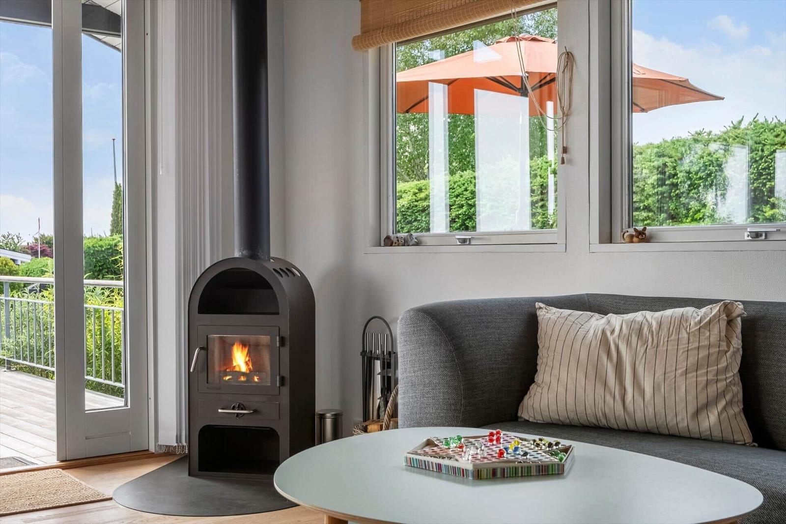 Ein Wohnzimmer mit Kamin, Sofa und Tisch. Durch Fenster ist ein Garten mit Sonnenschirm sichtbar.