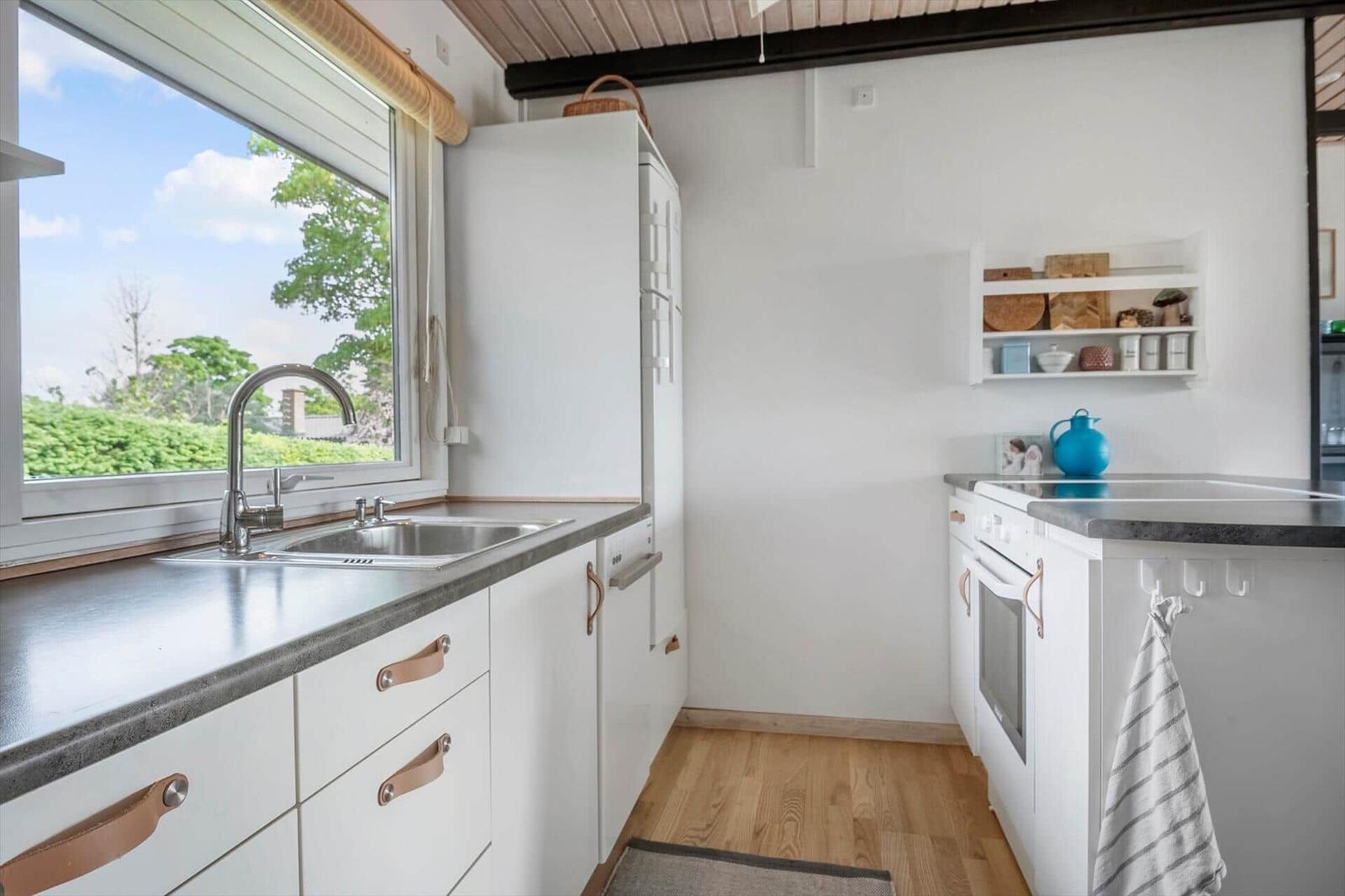 Küche mit Fenster, Spüle, Kühlschrank und Backofen. Holzboden und weiße Schränke.