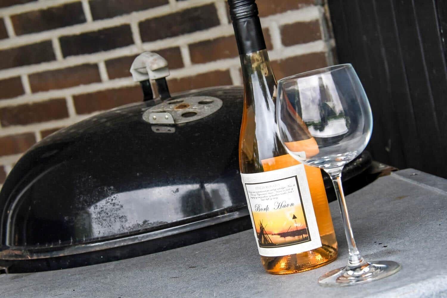 Eine Flasche Wein und ein Glas stehen neben einem Grill auf einer Terrasse.