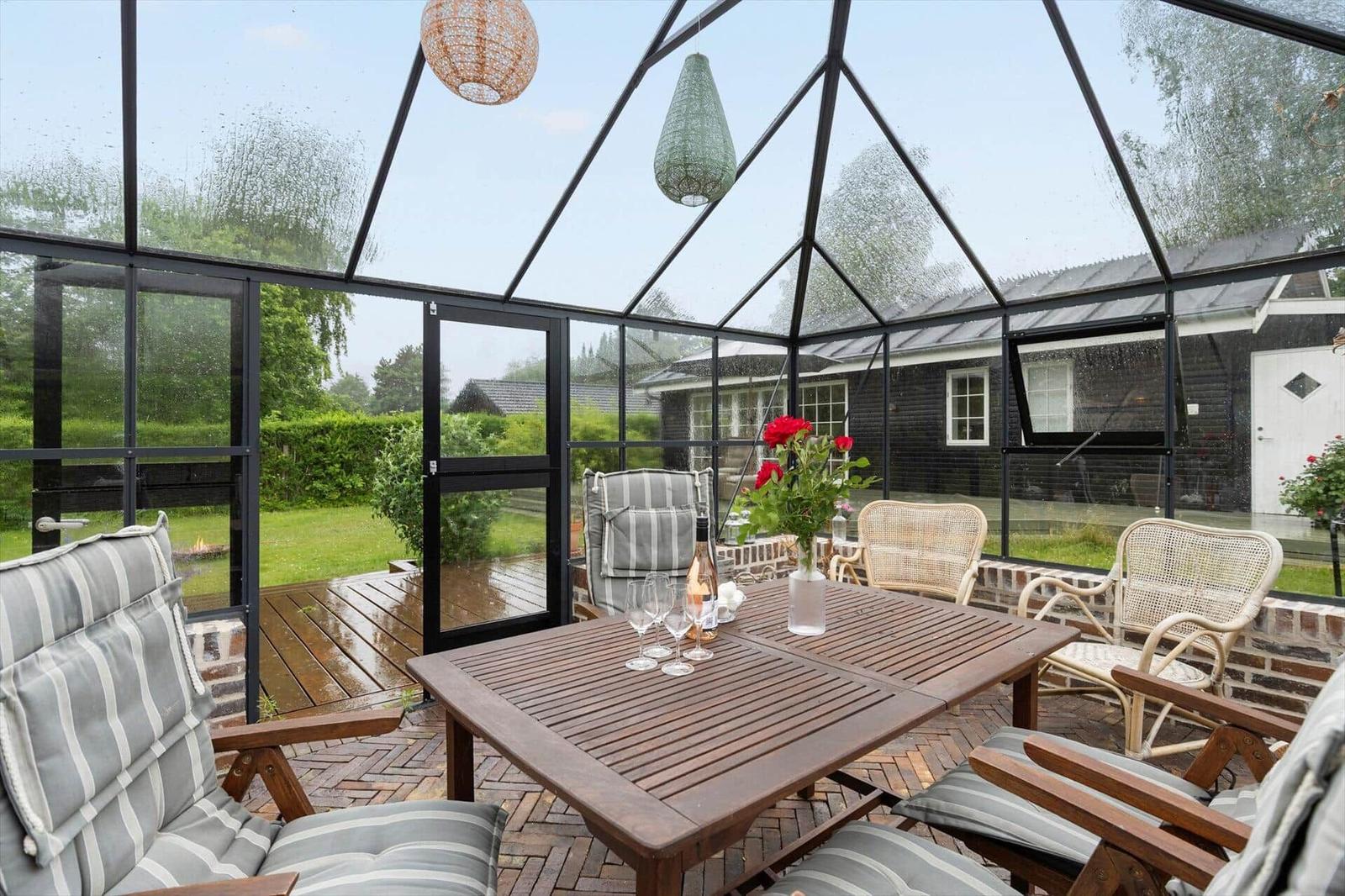 Glasveranda mit Holztisch, Stühlen und Blumen. Draußen ist ein Garten mit Rasen und Bäumen.