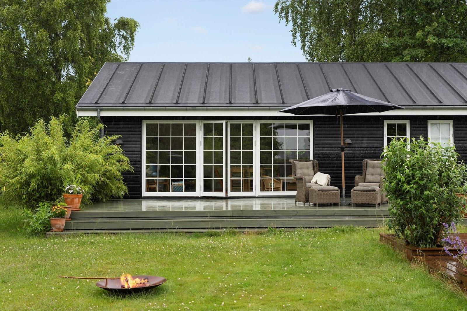 Schwarzes Haus mit Terrasse, Sitzgelegenheiten und Feuerkessel im Garten.