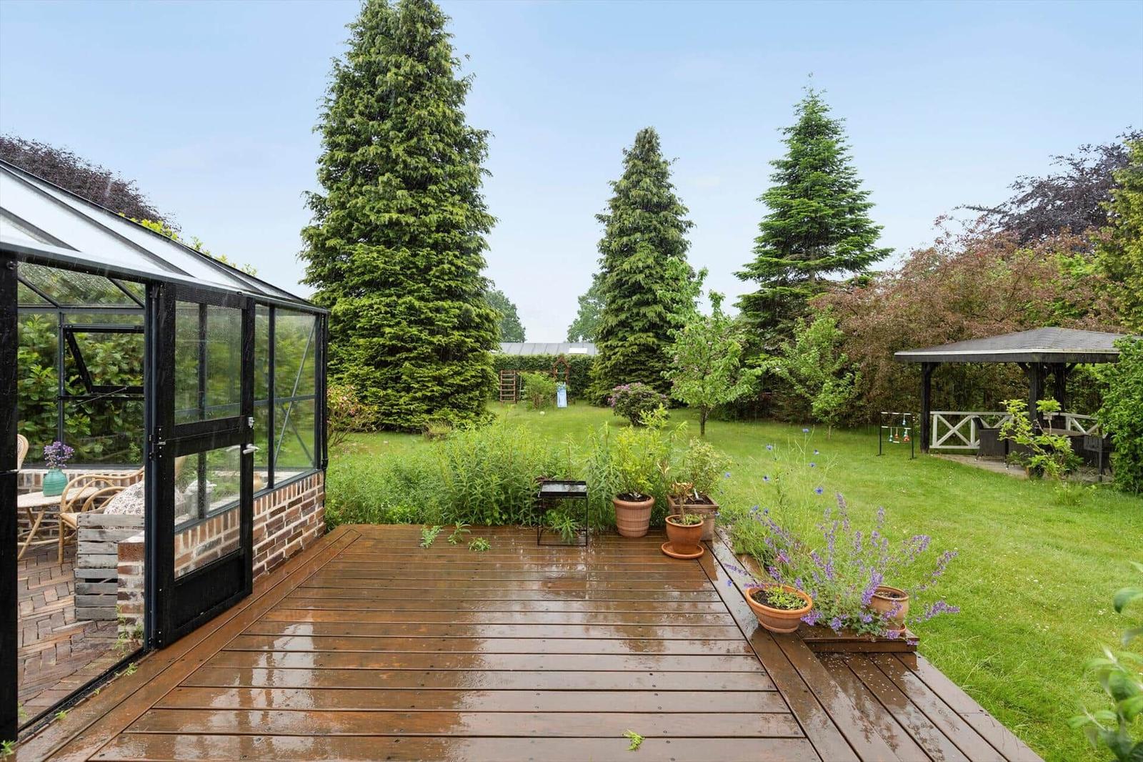 Holzterrasse mit Pflanzen, Gartenhaus und Pavillon im Hintergrund.