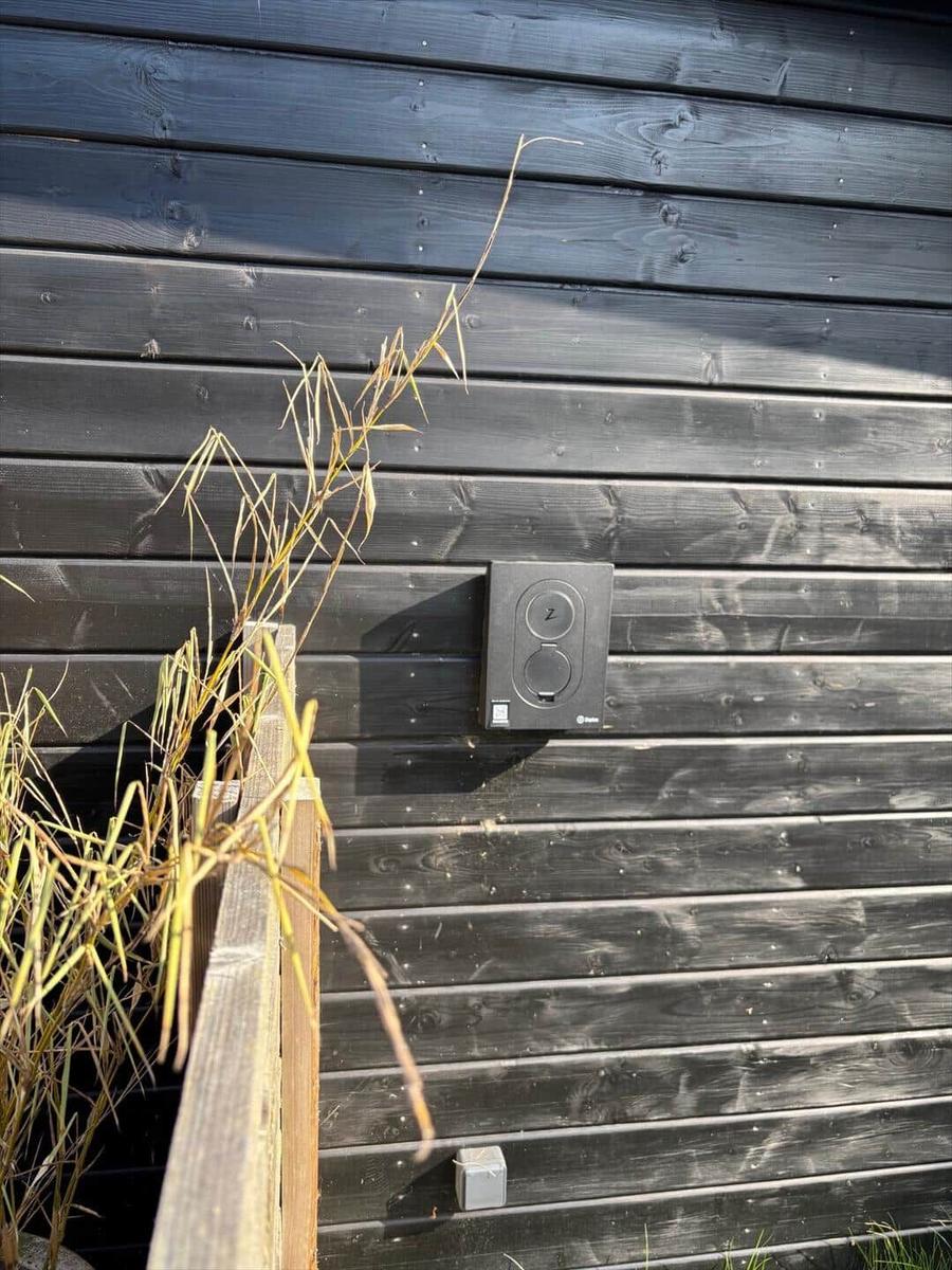 Black wooden wall with outlet and dry grass in the foreground.