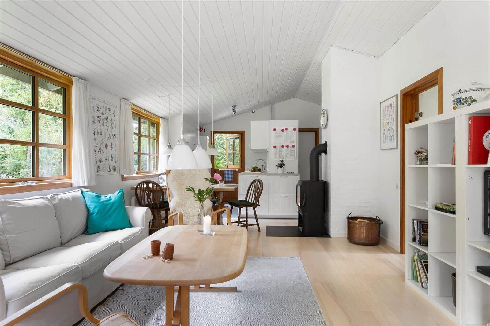 Wohnzimmer mit Sofa, Tisch und Küchenbereich. Holzdielen, weiße Wände und Fenster mit Holzrahmen.