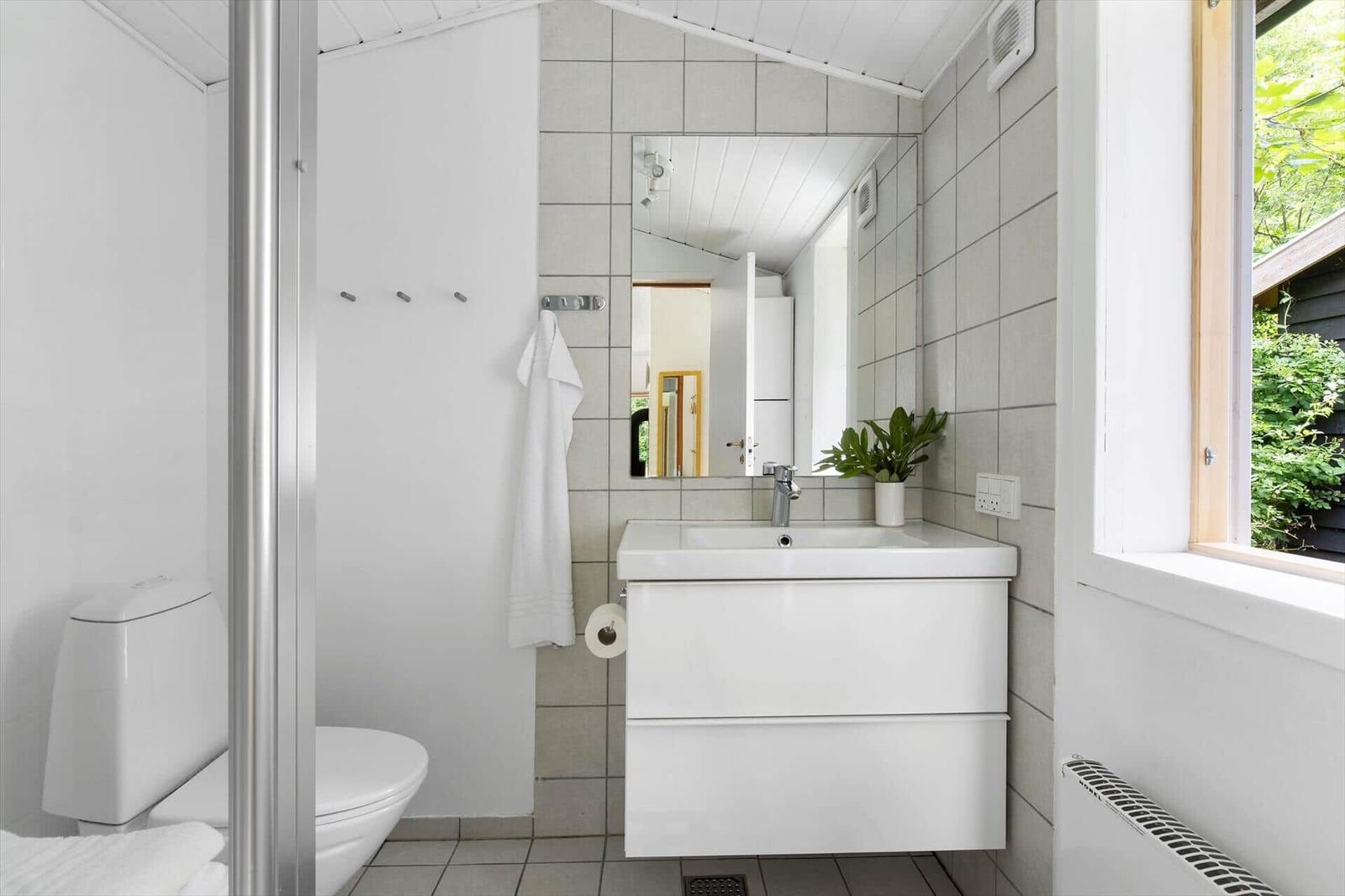 Badezimmer mit Waschbecken, Spiegel, Toilette und Dusche. Fenster mit Blick auf Grün.