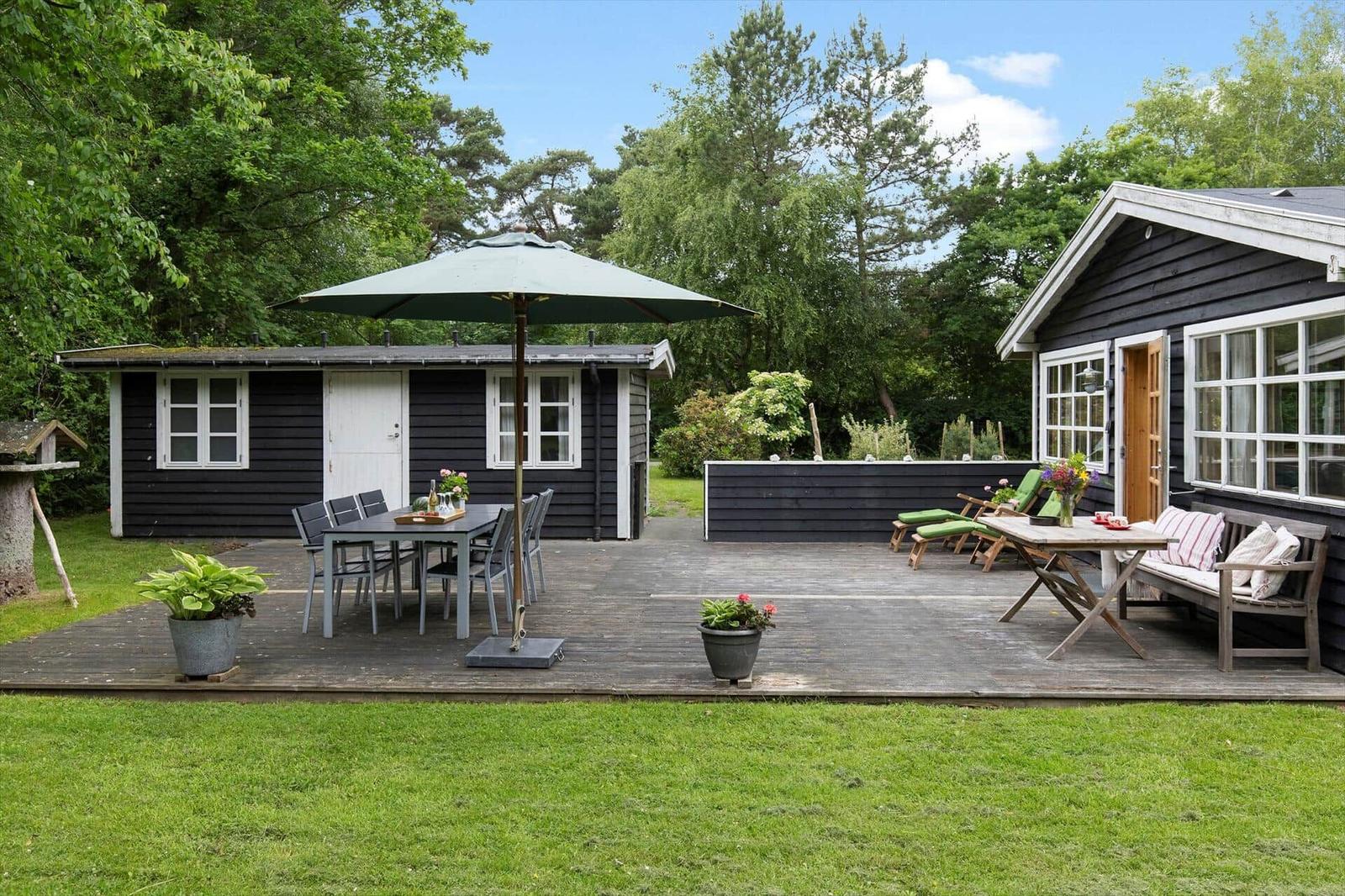 Helle Terrasse mit Tisch, Stühlen und Sonnenschirm. Schwarze Häuser mit grünem Garten.
