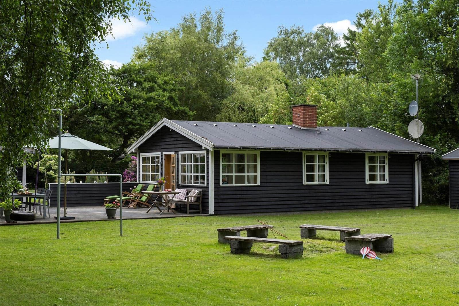 Schwarzes Holzhaus mit Terrasse, Garten und Bänken. Umgeben von Bäumen und Gras.