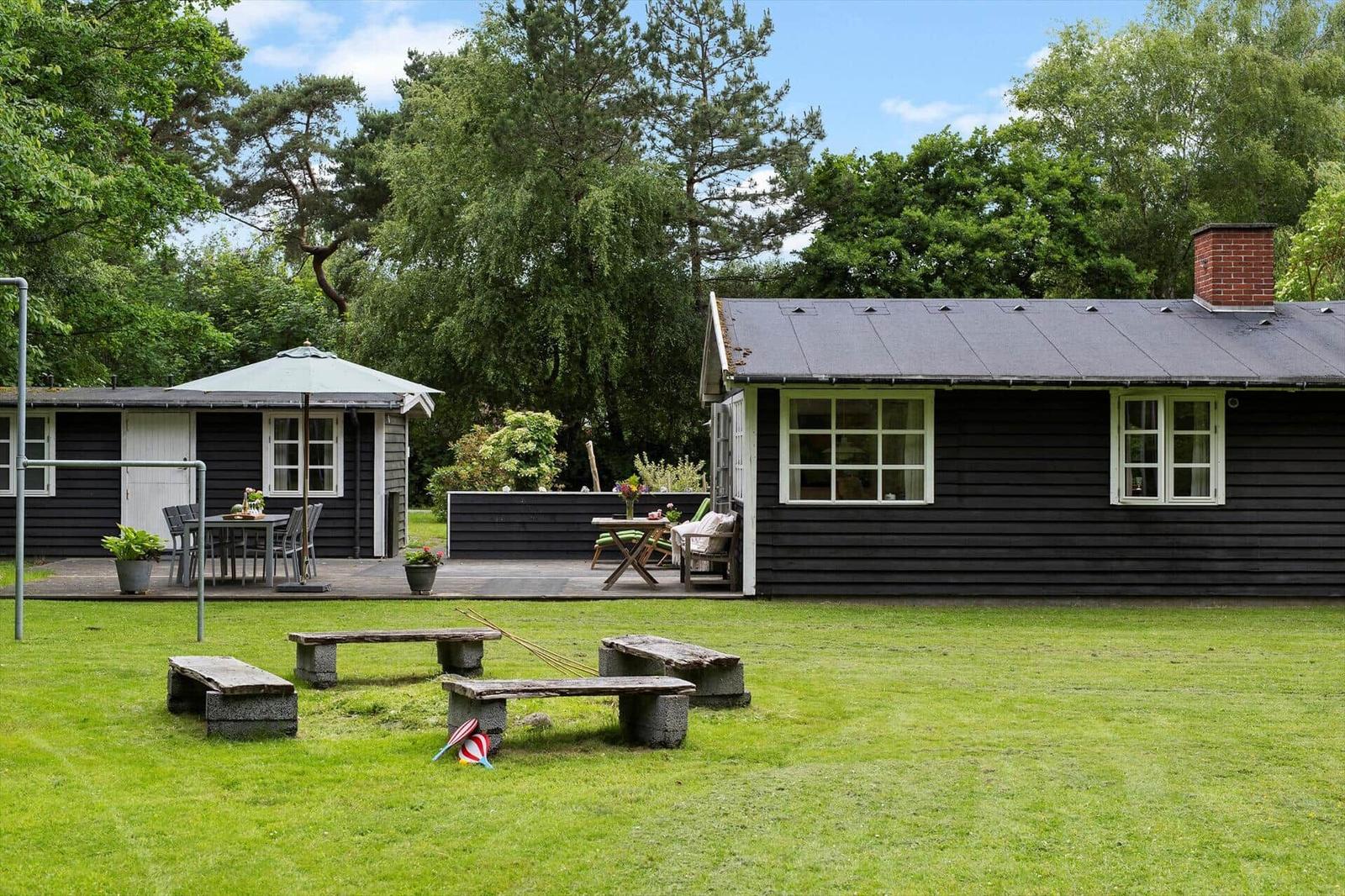Schwarze Holzhütte mit Garten, Tisch, Bänken und Sonnenschirm. Umgeben von Bäumen.