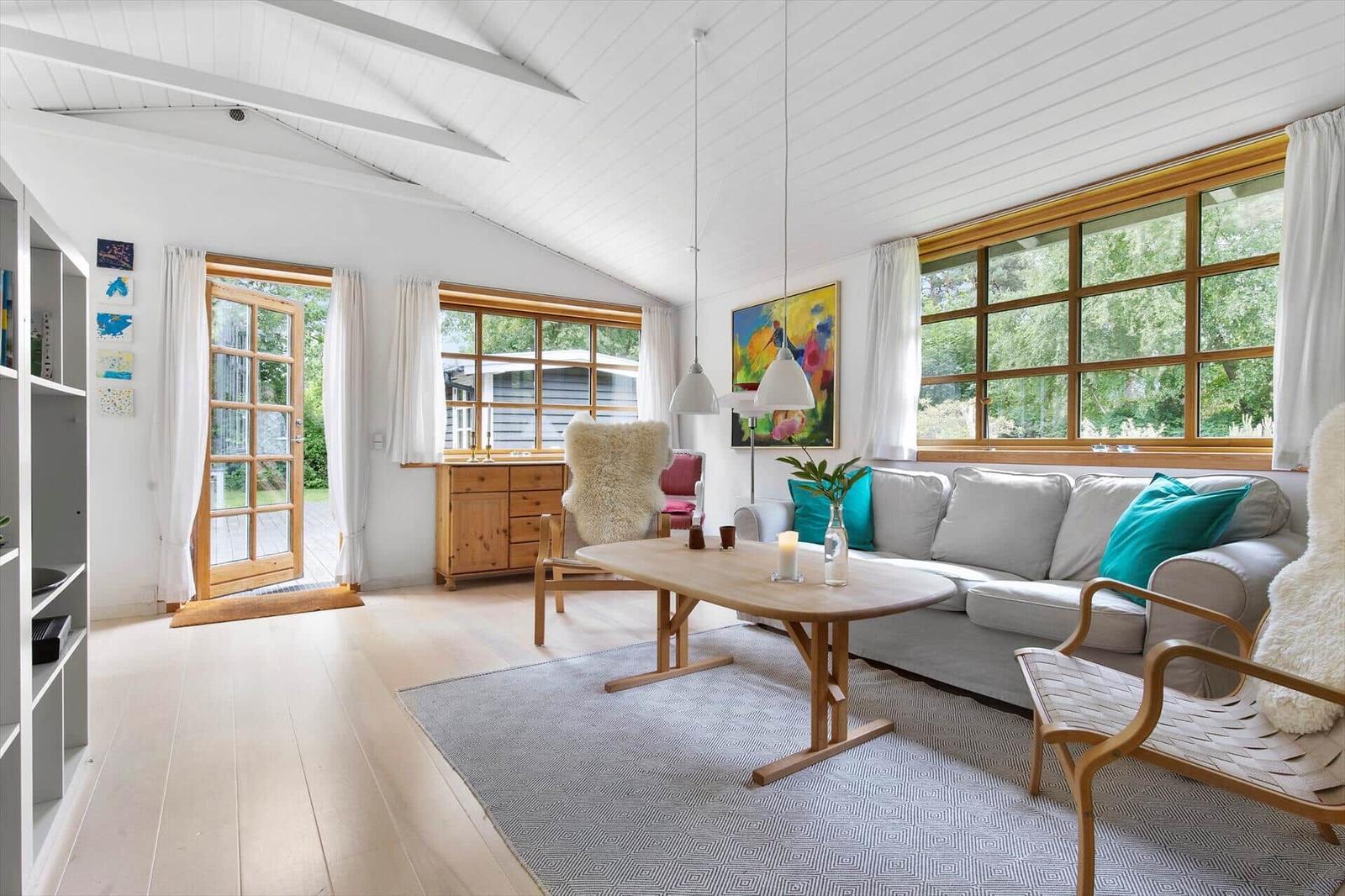 Wohnzimmer mit Sofa, Tisch und Stühlen. Große Fenster und Tür zur Außenterrasse.