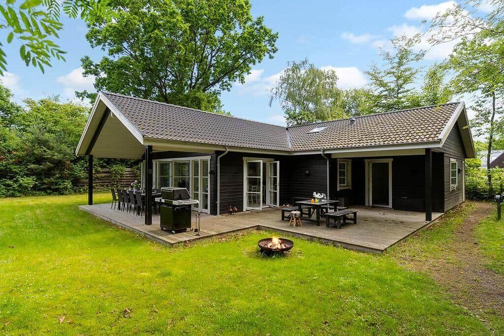 Ein Haus mit Terrasse, Grill und Feuerkorb im Garten. Umgeben von Bäumen und Gras.