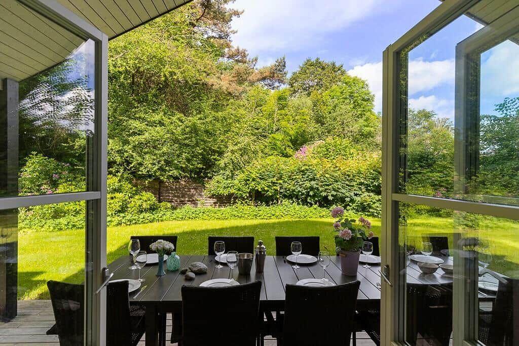 Gartenbereich mit Esstisch und Stühlen. Durchgehendes Glasfenster mit Blick auf grüne Landschaft.