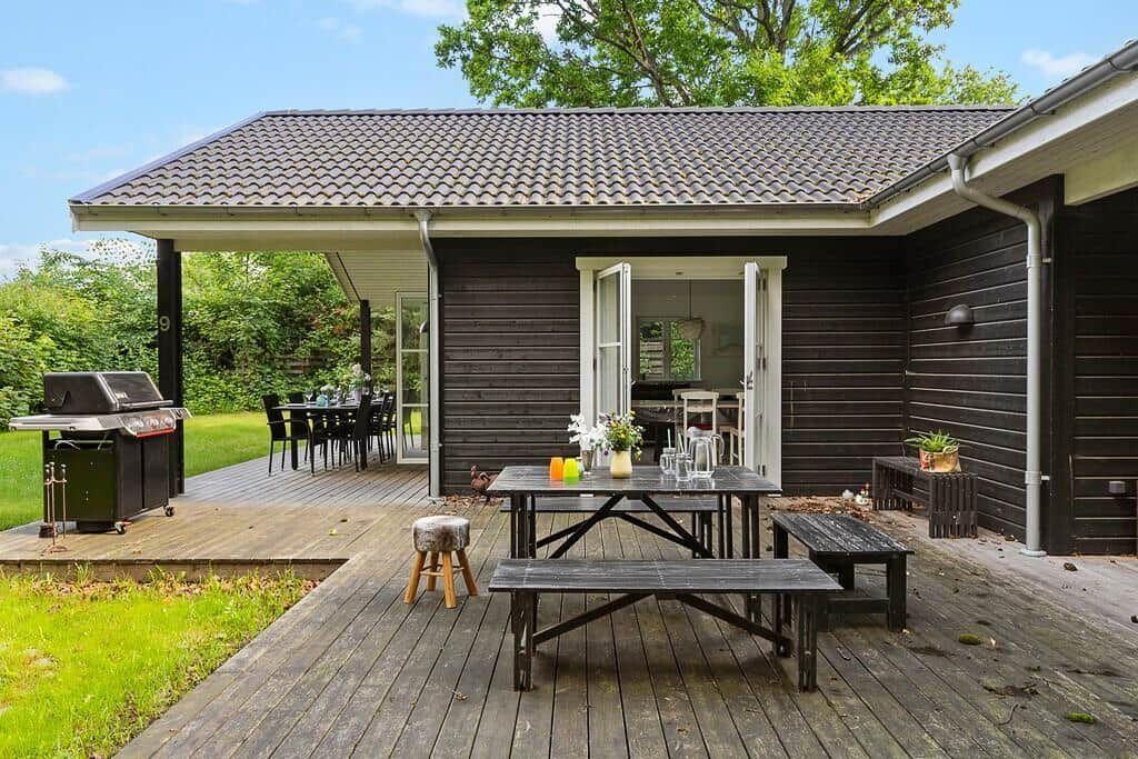 Terrasse mit Grill, Tisch und Bänken. Hintergrund: Haus mit offenen Türen.