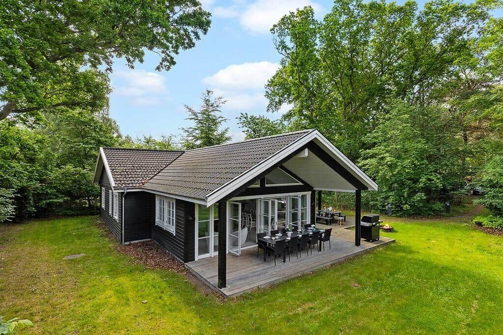 Schwarze Hütte mit Terrasse, Grill und Garten. Umgeben von Bäumen unter blauem Himmel.