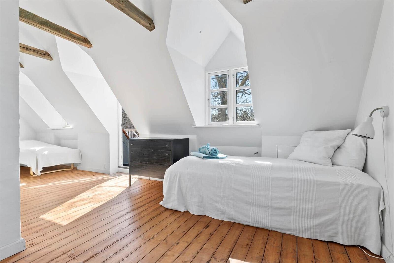 Schlafzimmer mit Dachschrägen, Holzboden und Fenster. Bett, Nachttisch und Lampe sind vorhanden.