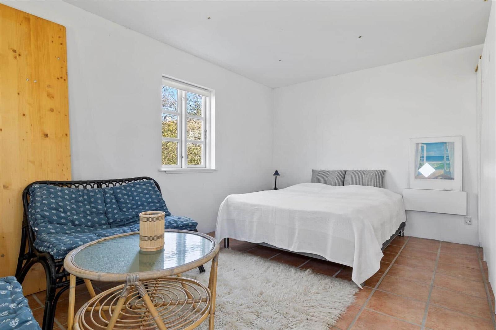 Schlafzimmer mit Bett, Fenster, Tisch und Sitzgelegenheit. Holzwand und Teppichboden.