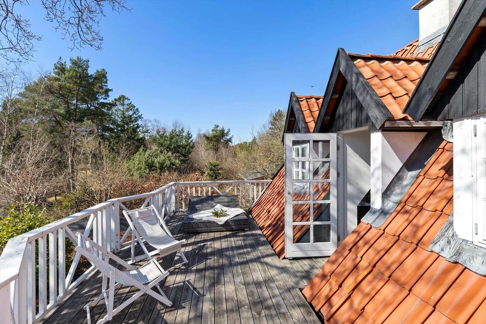 Terrasse mit Liege und Stühlen, Blick auf Wald und roten Dachfirst.