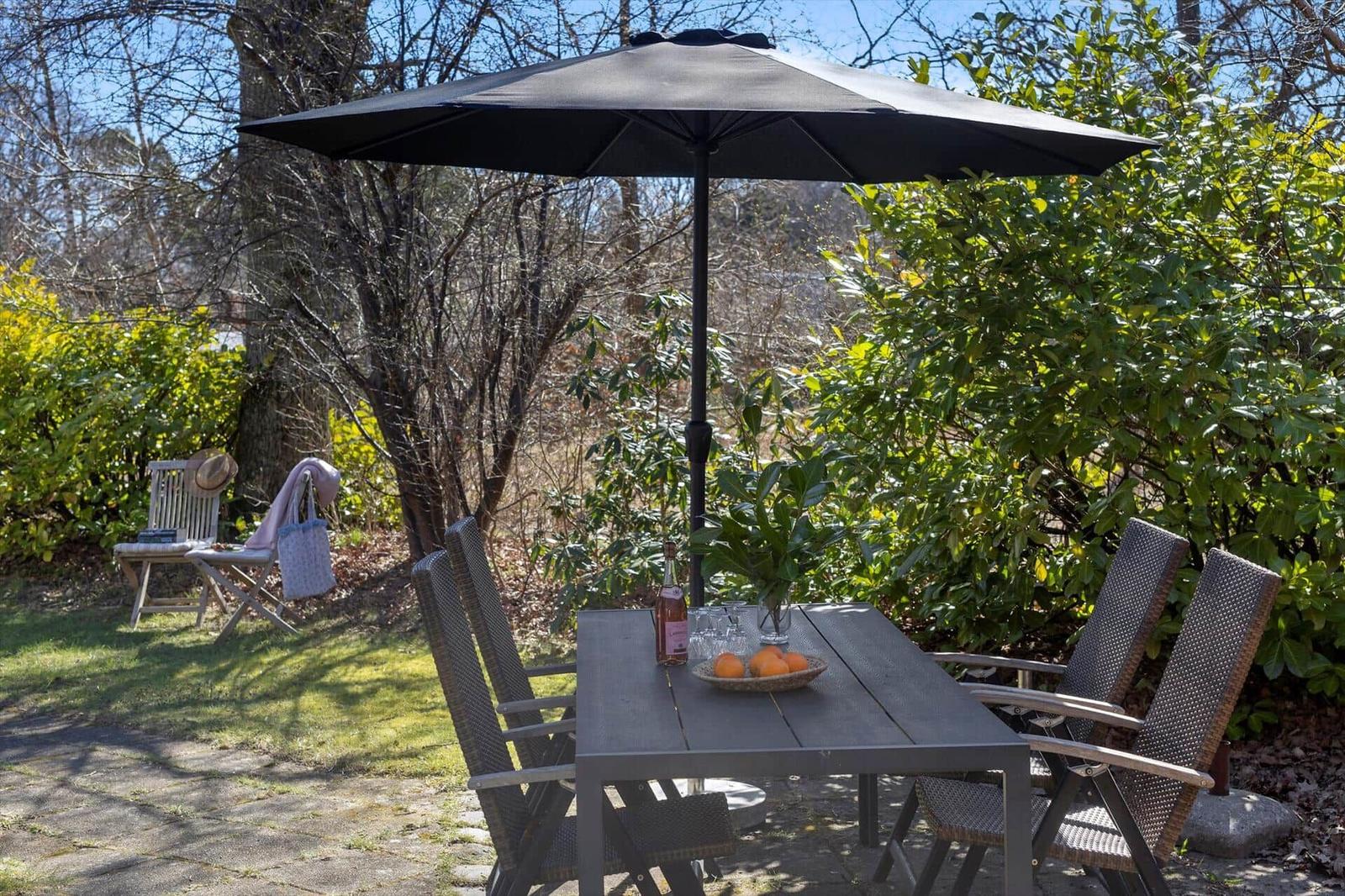 Terrasse mit Tisch, Stühlen und Sonnenschirm. Hintergrund mit Bäumen und Sträuchern.