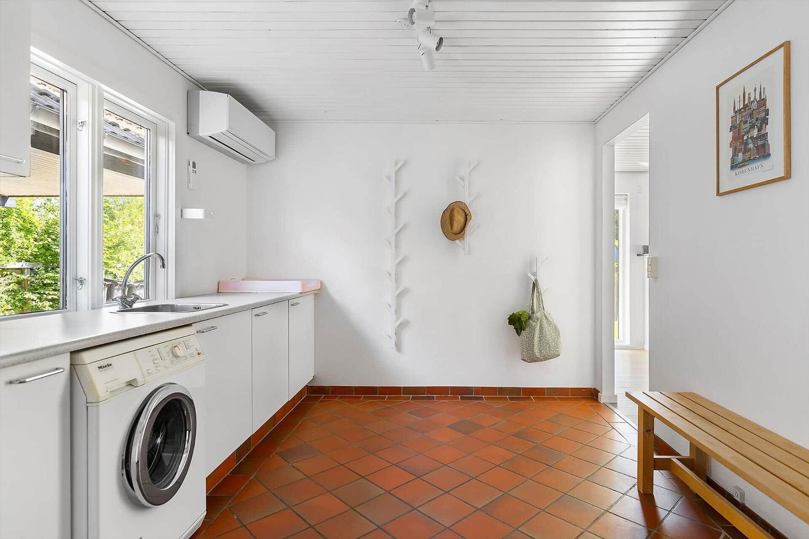 Küche mit Waschmaschine, Fenster und Holzbank. Weiße Wände, Terrakottboden und Haken.