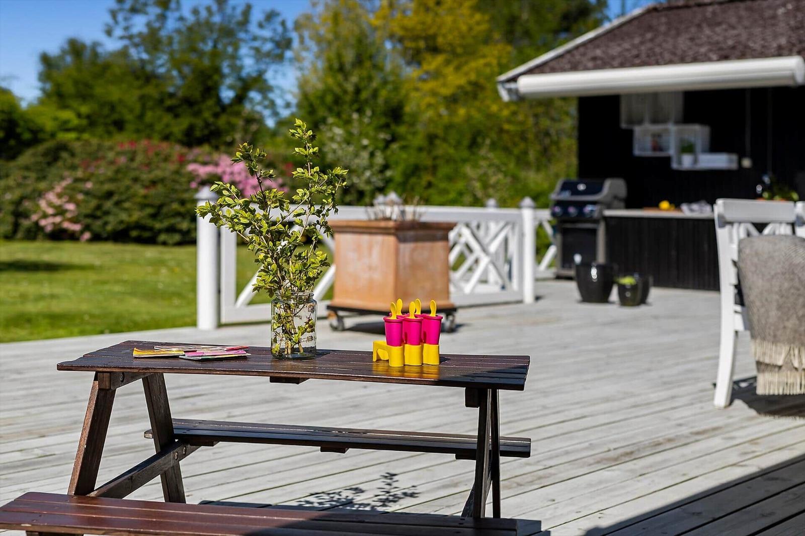 Holztisch mit Blumen und Eisbechern auf Terrasse. Grillhütte und Garten im Hintergrund.