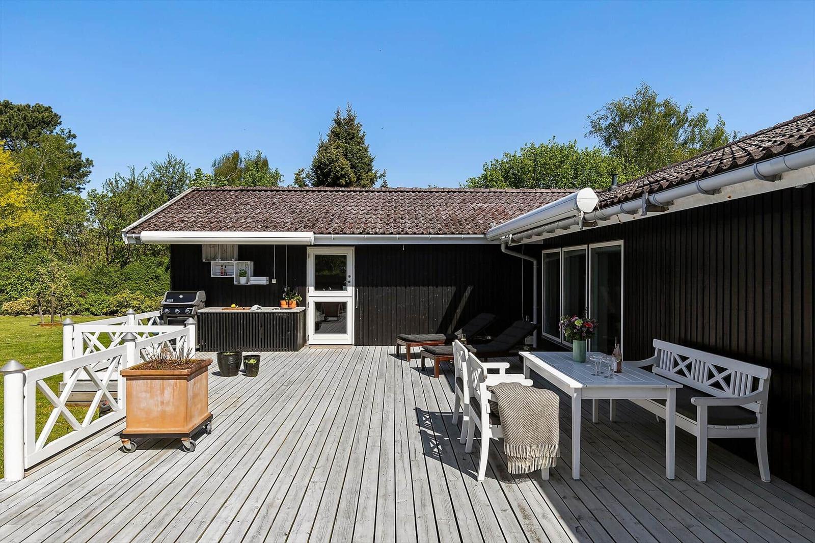 Terrasse mit Tisch, Bänken und Grill. Schwarze Fassade, weißer Zaun und Garten.