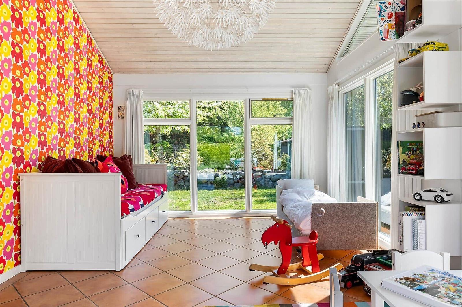 Kinderzimmer mit buntem Wandbehang, Schlafcouch, Spielzeug und großem Fensterblick auf Garten.