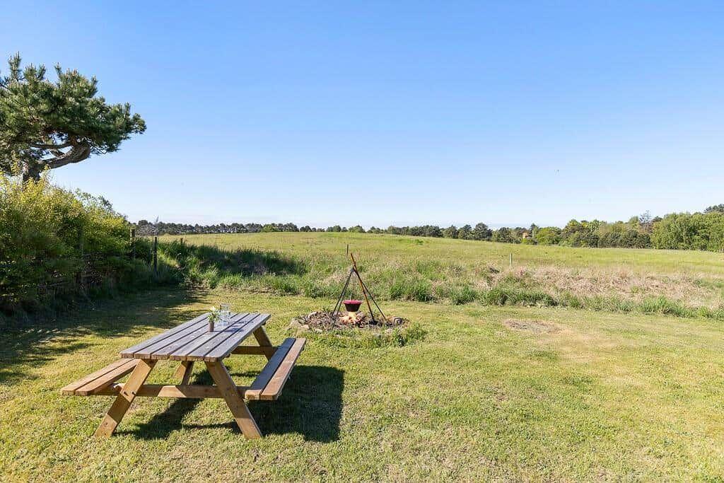 Holztisch und Grillplatz auf grünem Rasen mit Blick auf Feld und Bäume.