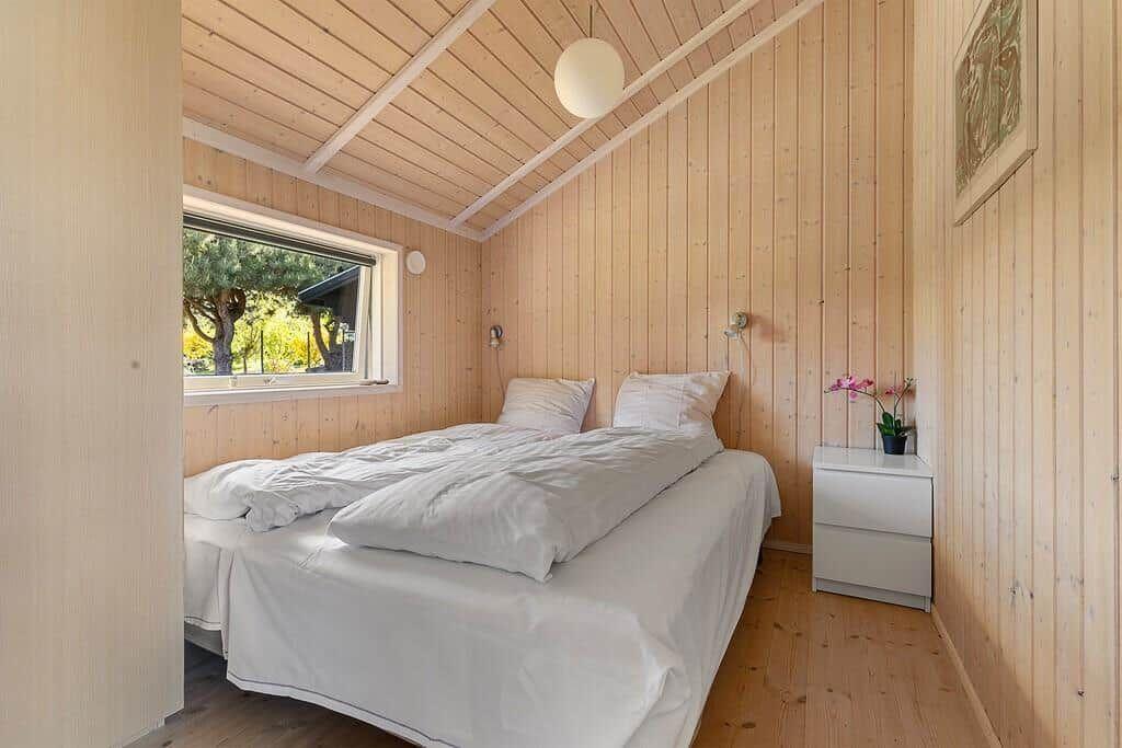 Schlafzimmer mit Bett, Fenster und Holzwandverkleidung. Neben dem Bett steht ein Nachttisch.