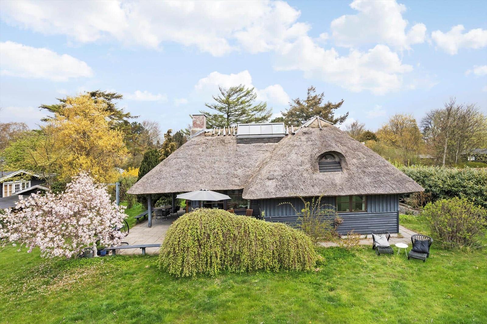 Haus mit Strohdach, Garten, Terrasse und Sitzgelegenheiten unter blauem Himmel.