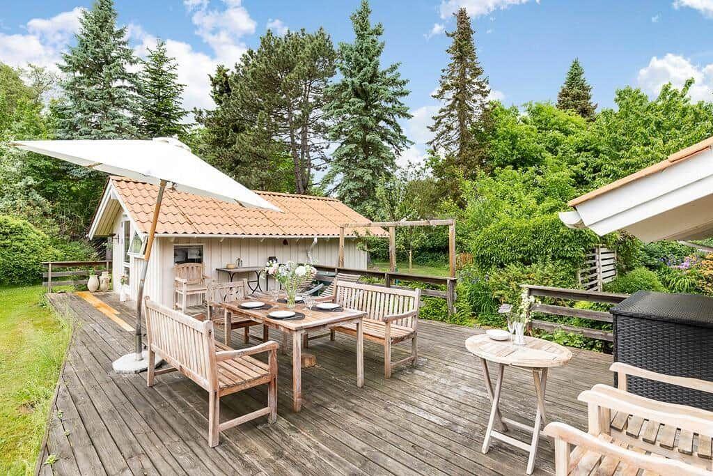 Holzterrasse mit Tisch, Bänken und Sonnenschirm. Hintergrund: Wald und Garten.