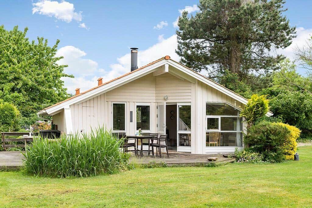 Ein weißes Haus mit Terrasse, Garten und Bäumen. Tisch und Stühle stehen draußen.