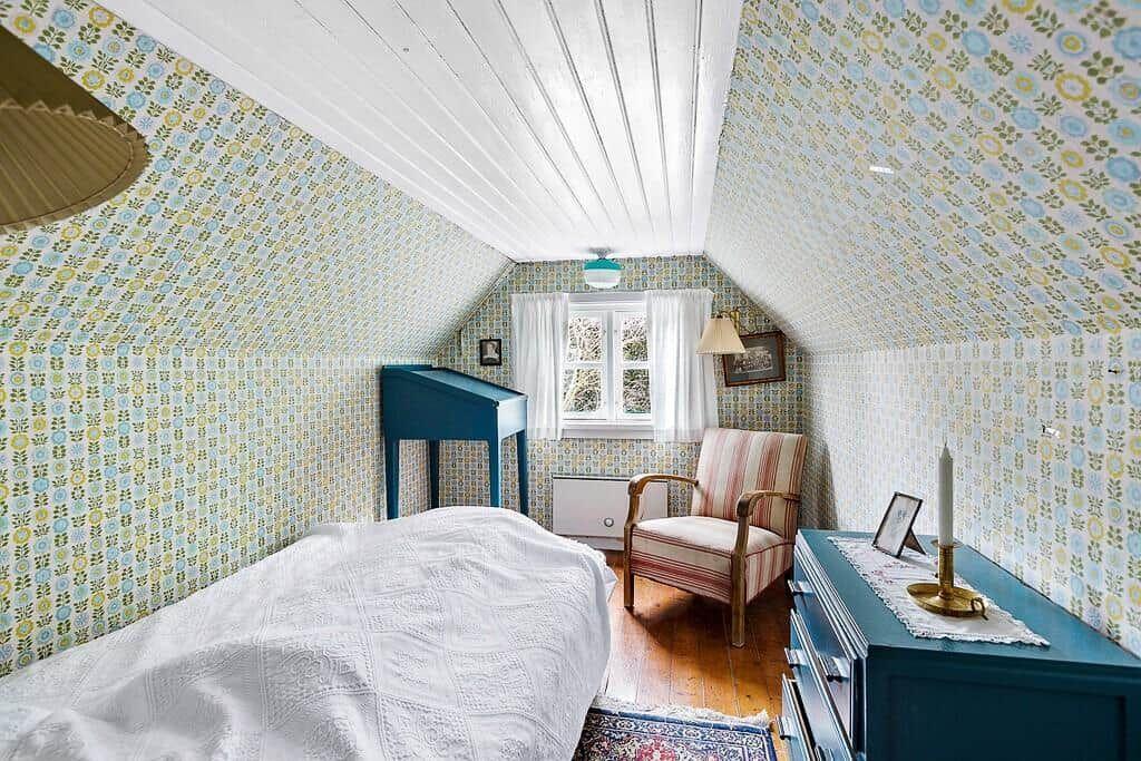 Schlafzimmer mit gemustertem Tapeten, Bett, Sessel und Schrank unter Dachschrägen.