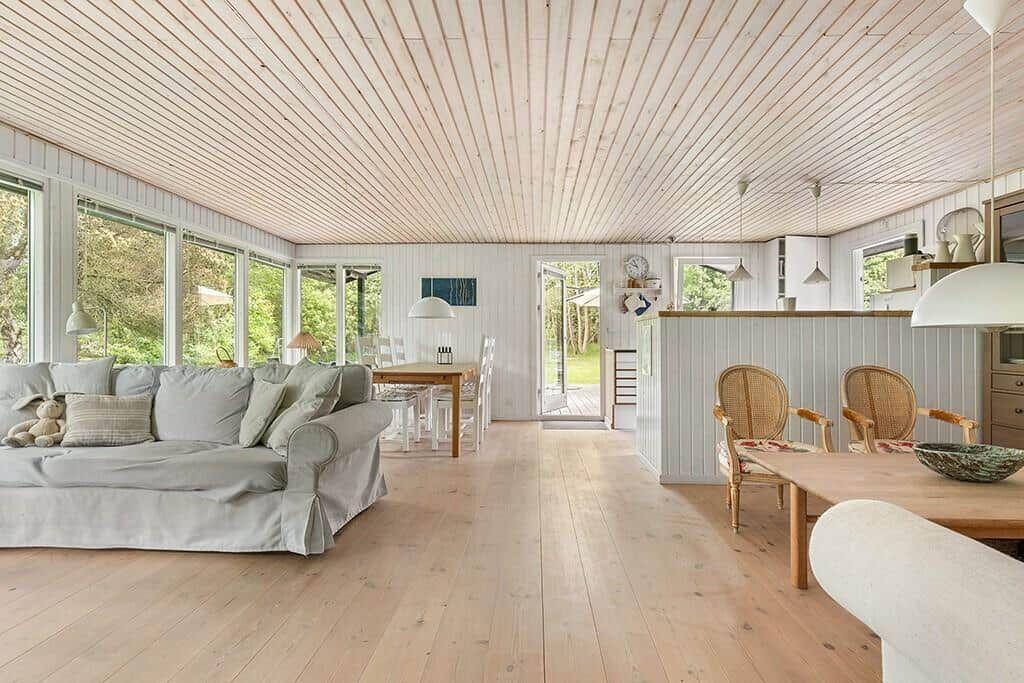Wohn- und Essbereich mit Holzboden, Sofa, Tisch und Stühlen. Große Fenster und Tür zur Außenterrasse.