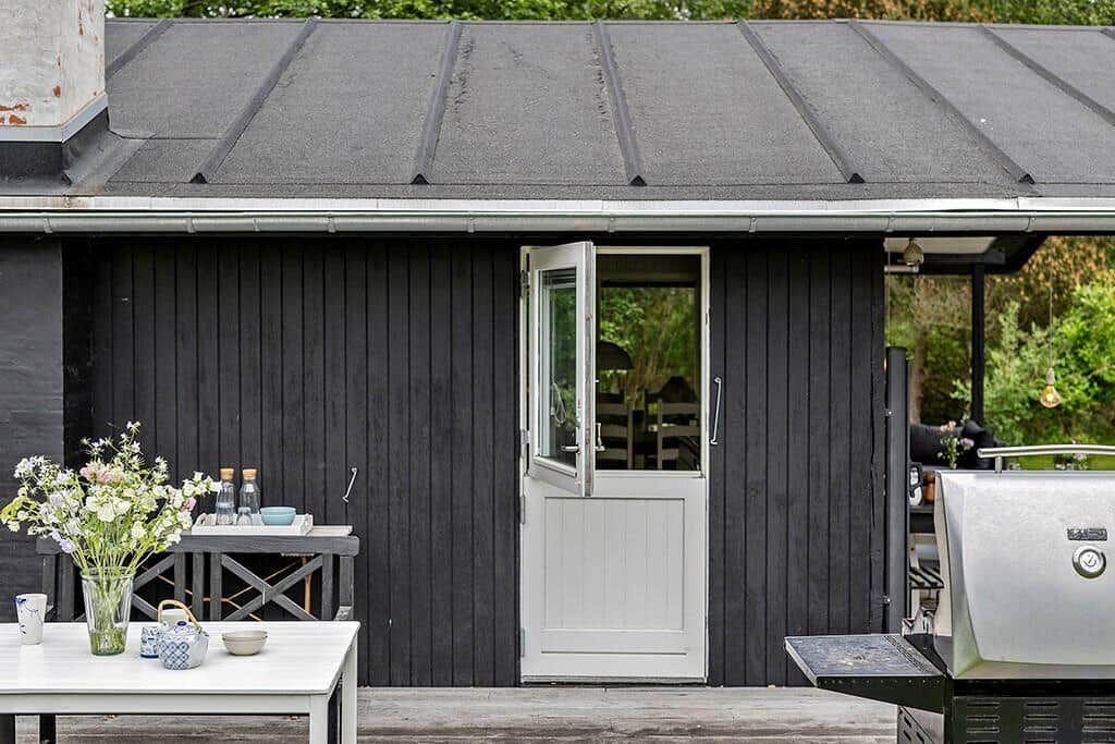 Eine schwarze Hütte mit weißer Tür, Terrasse mit Tisch, Blumen und Grill.