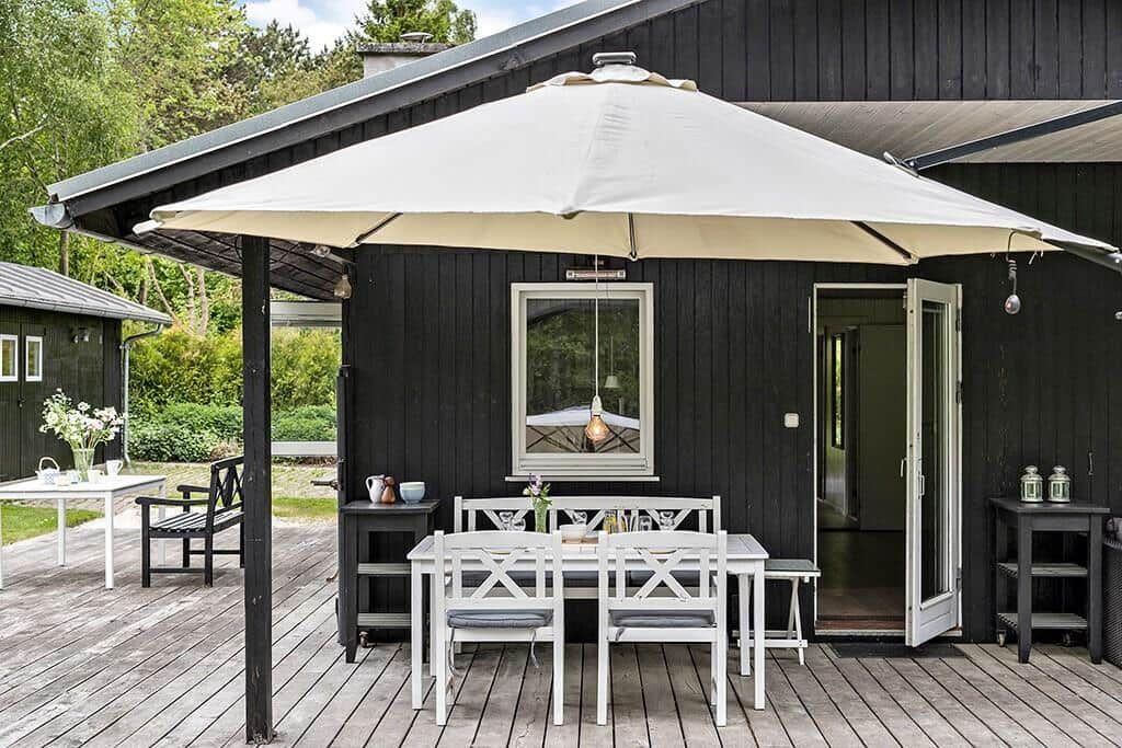 Terrasse mit weißem Tisch, Stühlen und Sonnenschirm. Schwarze Holzwände und offene Tür.