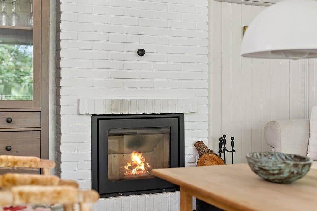 Kamin mit brennendem Feuer, weiß verputzte Mauer, Holztisch und Stuhl im Vordergrund.