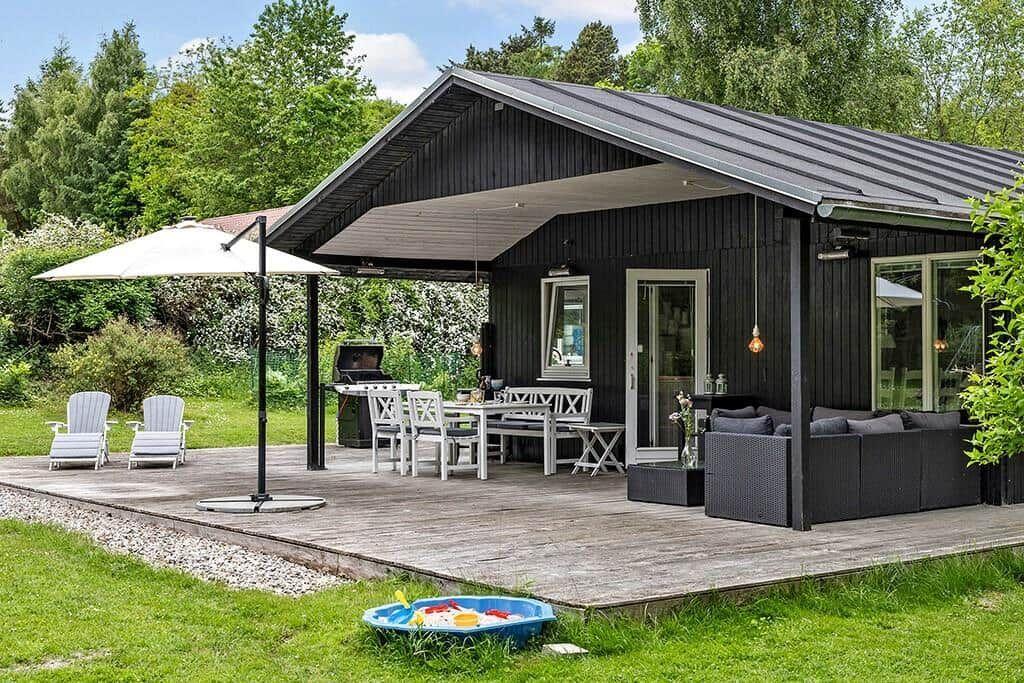 Schwarze Holzhütte mit Terrasse, Grill, Sitzgruppe und Kinderspielwasserbecken im Garten.