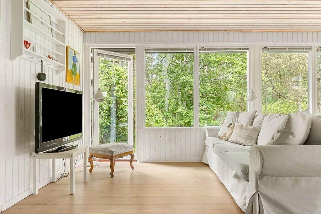 Wohnzimmer mit Sofa, Fernseher und Blick auf grüne Bäume durch große Fenster.