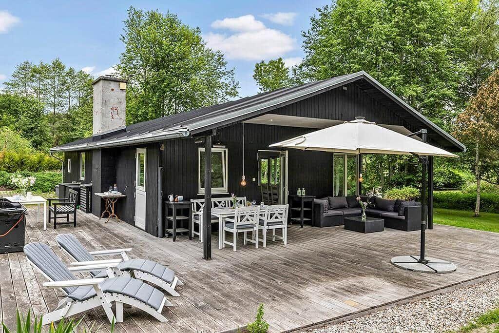 Schwarzes Haus mit Holzterrasse, Gartenmöbeln und Sonnenschutz. Umgeben von Bäumen und Grün.