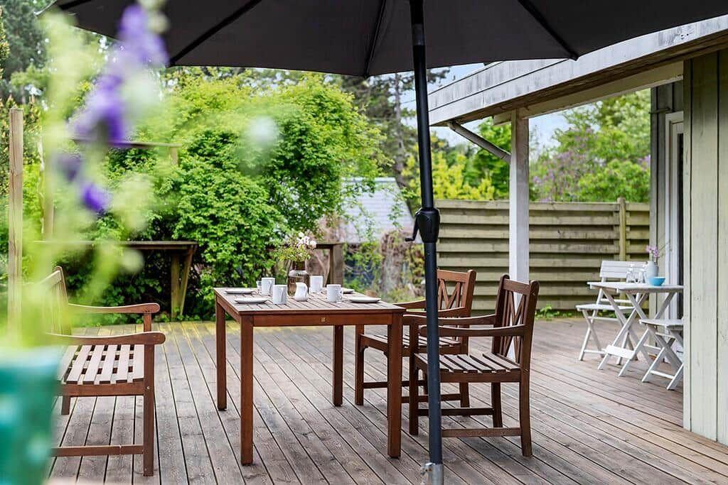 Terrasse mit Tisch, Stühlen und Sonnenschutz. Holzdielen und grüne Umgebung.