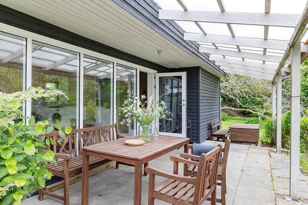 Terrasse mit Holztisch, Stühlen und Pergola. Durchgehendes Glasfensterfront. Grünflächen im Hintergrund.