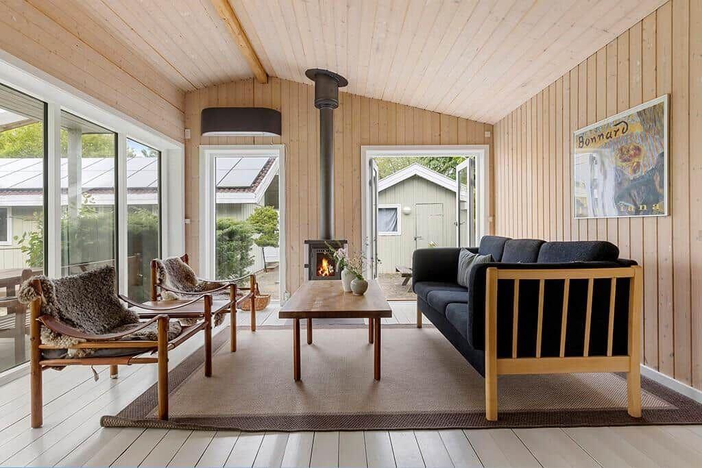 Wohnraum mit Sofa, Holztisch und Kaminofen. Holzwände und große Fenster.