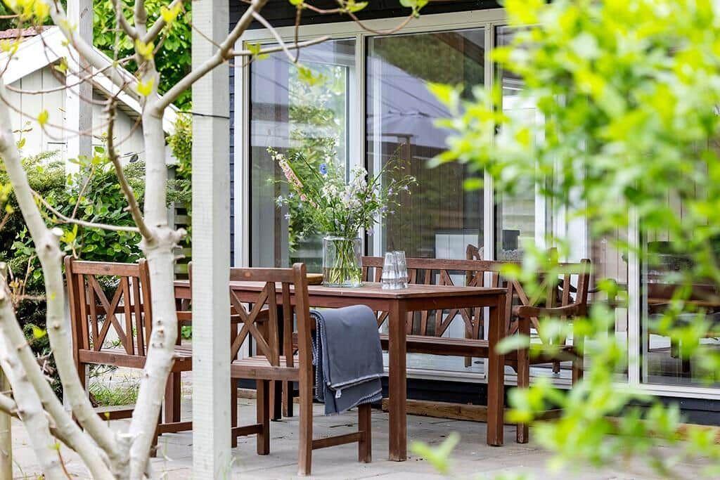Holztisch mit Stühlen und Blumen auf einer Terrasse vor einem Haus mit Fenstern.
