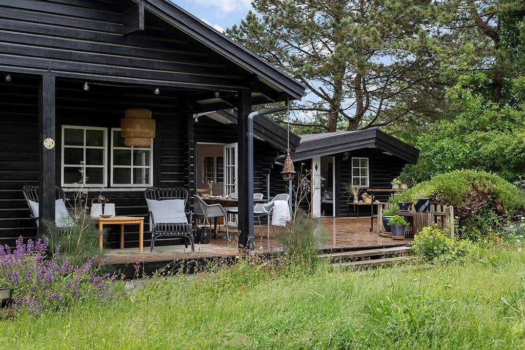 Schwarzes Holzhaus mit Terrasse, Garten und Bäumen. Möbel und Pflanzen sind sichtbar.