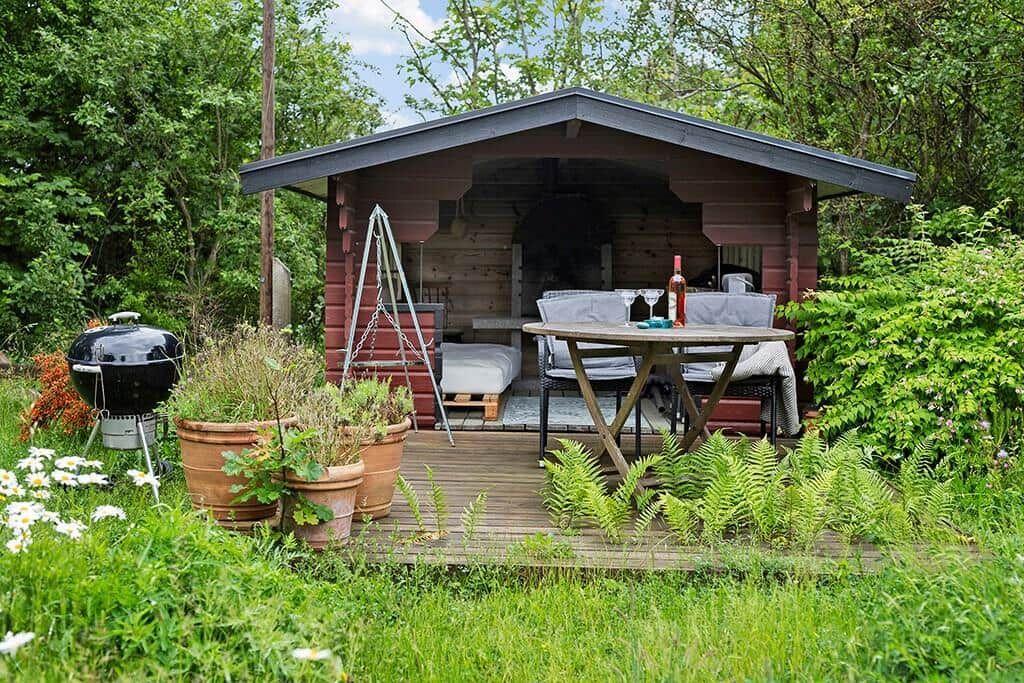 Hölzerner Gartenbau mit Tisch, Stühlen und Grill. Pflanzen und Gras umgeben die Terrasse.