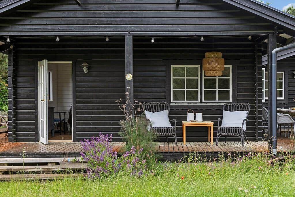 Schwarze Holzhütte mit Terrasse, zwei Stühlen und Tisch. Tür offen, Blick ins Innere.